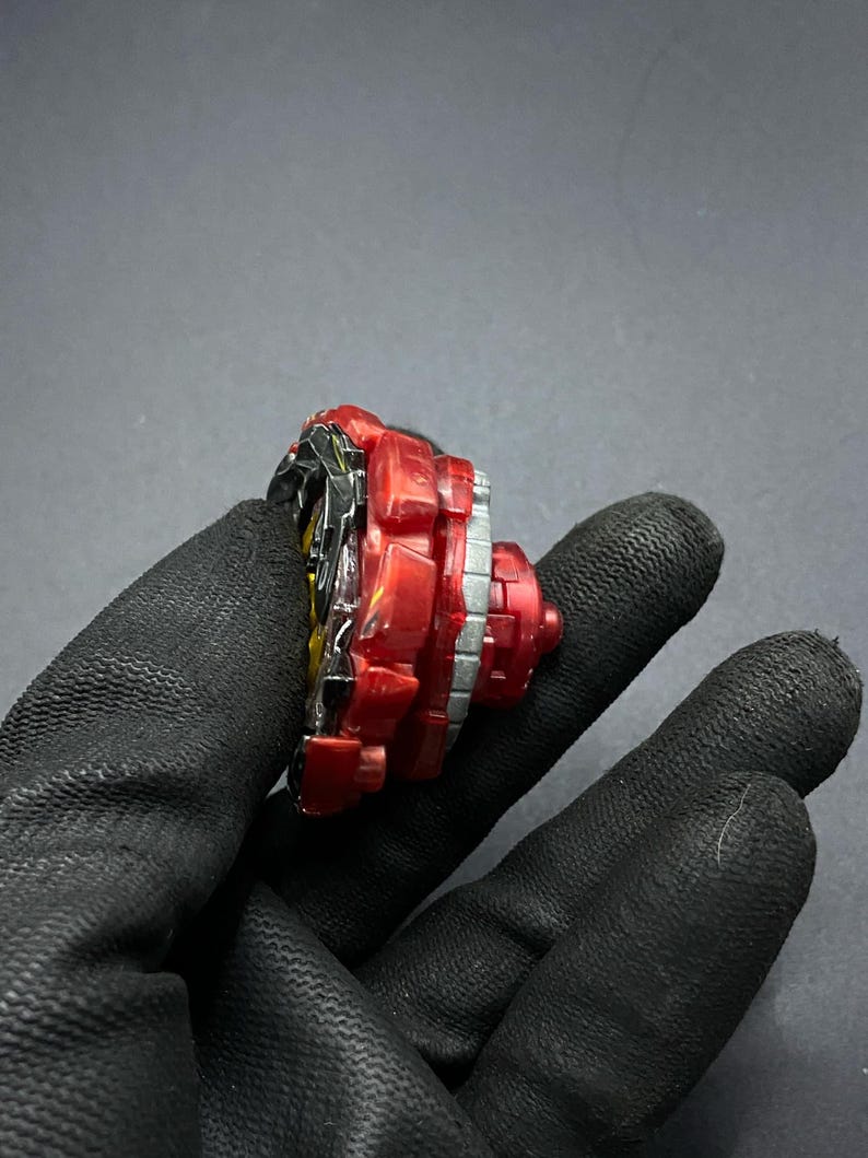 K&ouml;nnte beinhalten: Ein rotes und schwarzes Beyblade mit einer silbernen und wei&szlig;en Schicht. Das Beyblade wird in einer behandschuhten Hand gehalten.