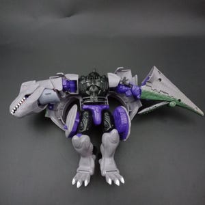 May include: A gray and purple action figure in a dinosaur-like form, with a detailed design. The figure has a dinosaur head, wings, and legs, with purple accents on the body. The figure is posed against a dark background.