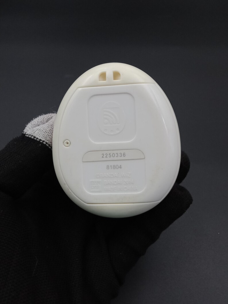 Puede incluir: Un dispositivo electr&oacute;nico blanco de forma ovalada con un panel rectangular y dos peque&ntilde;as protuberancias en la parte superior. El dispositivo tiene los n&uacute;meros "2250336" y "81804" impresos. El dispositivo es sostenido por una mano enguantada.