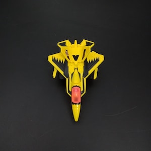 May include: A yellow toy jet with black accents and a red cockpit. The toy has a pointed nose and wings with jagged edges. The jet is designed with a shark-like mouth on the front.