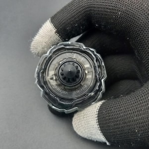 Pu&ograve; includere: Un giocattolo trottola trasparente e nero con un centro a forma di ingranaggio e un anello esterno trasparente. Il giocattolo &egrave; tenuto da una mano guantata su uno sfondo grigio. I testi "TOMY" e "VIETNAM" sono visibili sul giocattolo.