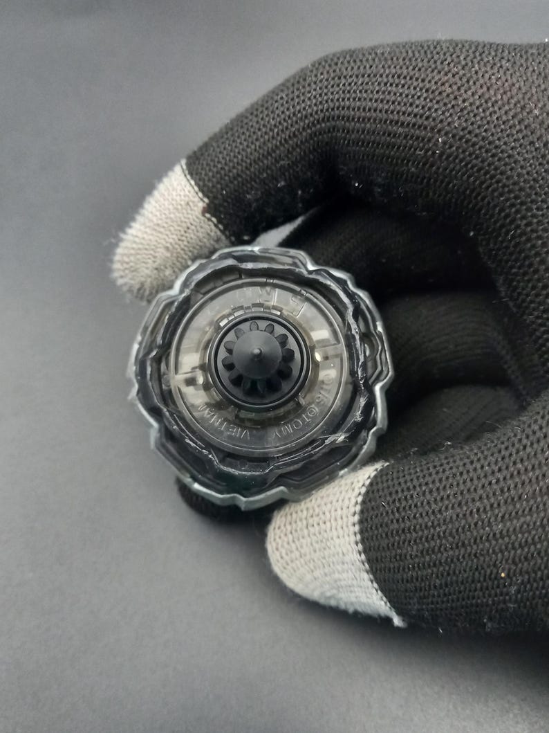 Pu&ograve; includere: Un giocattolo trottola trasparente e nero con un centro a forma di ingranaggio e un anello esterno trasparente. Il giocattolo &egrave; tenuto da una mano guantata su uno sfondo grigio. I testi "TOMY" e "VIETNAM" sono visibili sul giocattolo.
