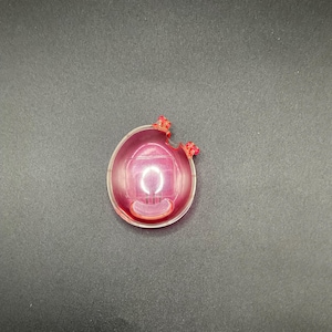 May include: A clear, oval-shaped, pink plastic object with two red protrusions on top. The object has a small, rectangular opening at the bottom and a curved, white design inside. The object is on a dark gray surface.
