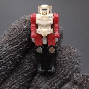 May include: A small, vintage action figure of a robot with a white chest and head, red arms and legs, and black feet. The robot has blue eyes and is standing upright. The figure is a collectible toy.