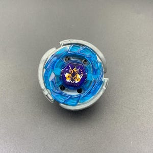 Puede incluir: Un juguete de peonza circular con un anillo exterior de metal plateado y una sección central de plástico azul translúcido. En el centro, un perno hexagonal morado muestra una ilustración gráfica de una cabeza de caballo amarilla y blanca.