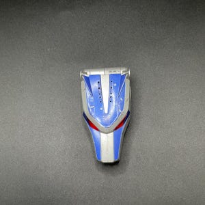 Peut inclure: Une tête de robot jouet bleue et argentée avec des détails d'yeux rouges. Le jouet a un design futuriste avec un dessus argenté et des accents bleus. La tête de robot est en plastique.