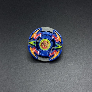 May include: A circular blue and grey spinning top toy featuring four curved arms with orange and green accents. The center of the toy displays a circular sticker with a red and yellow graphic illustration of a dragon-like creature on a yellow background.