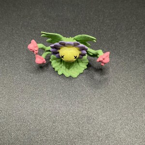 以下が含まれることがあります： 黄色い中心、紫色の花びら、緑の葉を持つ、手作りの花の置物。 花には、目のための2つの小さな黒いXと、側面から伸びるピンク色の花のつぼみがあります。 置物は柔らかく柔軟な素材で作られています。