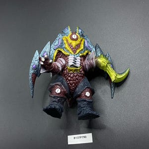 以下が含まれることがあります： 緑色の刃を持った赤、黒、黄色のモンスターのおもちゃ。モンスターは、大きな頭と片方の目、鋭い歯の口を持っています。おもちゃは、腕を伸ばし、足を曲げたポーズをしています。おもちゃには「B137P790」という番号が付けられています。