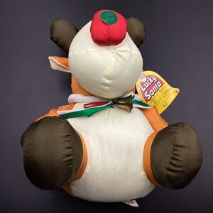 May include: A plush reindeer toy with a red hat, brown antlers, and a white body. The reindeer is wearing a green and white scarf and has a bell around its neck. The toy has a tag that says "Lucky Santa".