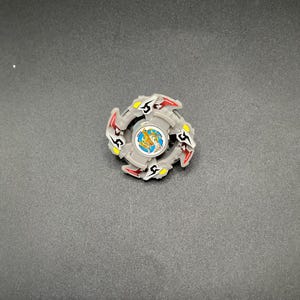 May include: A grey and red spinning top toy with a circular design. The toy has a central emblem with a gold and blue image. The top has red and yellow accents against a dark grey background.