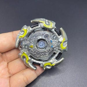 Puede incluir: Un juguete plateado y amarillo con un diseño circular. El juguete tiene una sección central transparente con un centro azul y está rodeado de detalles plateados y amarillos. El juguete se sostiene en una mano.