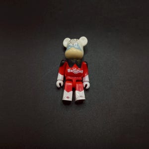May include: A small, red and white collectible figure with a bear-like head and a stylized outfit. The figure has a white head with bear ears, a tan face, and a red body with white accents. The figure is standing on a dark surface.