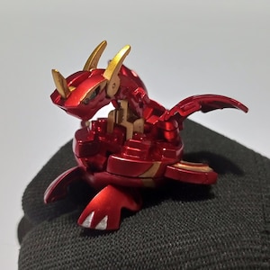Peut inclure: Une figurine de jouet en forme de dragon rouge métallique avec des cornes et des accents dorés. La créature présente un corps segmenté, des ailes angulaires pointues et des griffes avec des détails argentés sur les pieds, en position alerte.