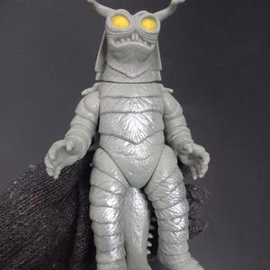 May include: A grey monster action figure with yellow eyes and horns. The figure has a textured, scaled appearance and is posed with its arms slightly outstretched. The figure is held by a dark grey glove.