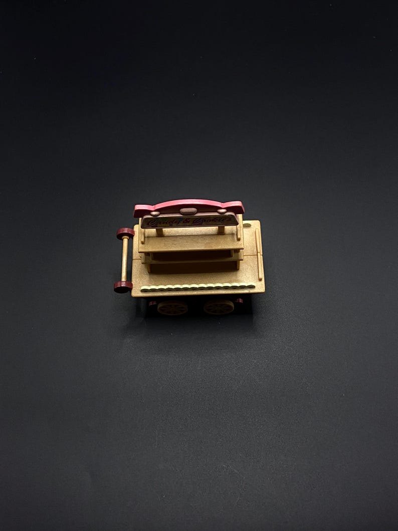May include: A miniature wooden cart with a red and tan color scheme. The cart has two shelves and four wheels. The top of the cart has a red sign that reads "Candy & Popcorn".