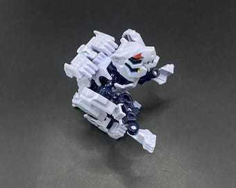 May include: A small, white and dark blue robotic toy figure with articulated joints. The toy has a detailed design with visible mechanical components and red accents on the head. The figure is posed in a dynamic, crouching position.