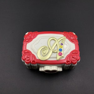 Pode incluir: Uma caixa de bugigangas retangular decorativa com um design vermelho e branco. A caixa apresenta uma letra "A" dourada adornada com pedras preciosas coloridas. As bordas são vermelhas com detalhes florais, e o centro é branco.
