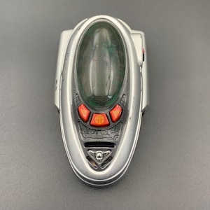 May include: A silver, oval-shaped electronic toy device featuring a dark green, translucent, dome-like center piece. It includes three bright orange buttons at the base of the dome, with a small circular emblem on the center button and black detailing.
