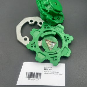 May include: Green Beyblade toy with a silver metal piece and a white label with text and a barcode. The toy is in pieces, with a circular design and a top piece. The label includes the text "Beyblade Draciel F Green".