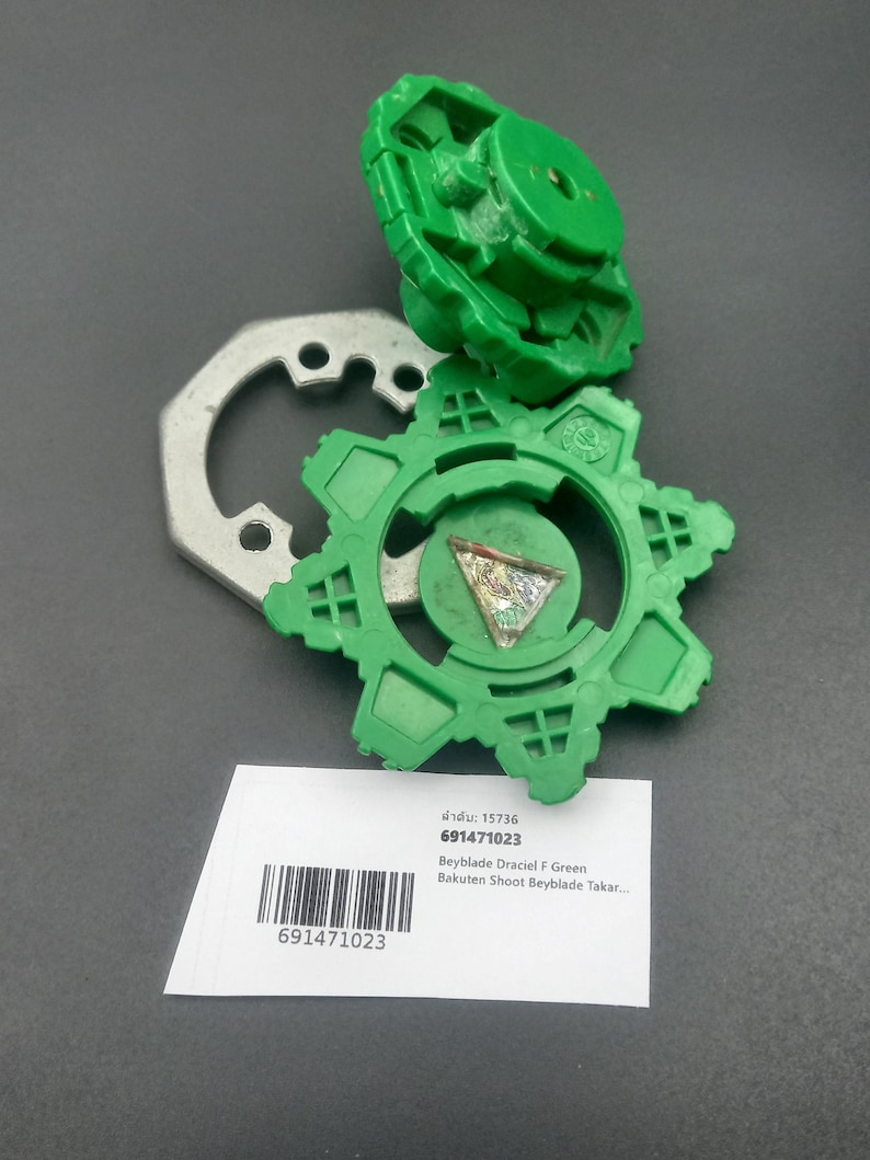 May include: Green Beyblade toy with a silver metal piece and a white label with text and a barcode. The toy is in pieces, with a circular design and a top piece. The label includes the text "Beyblade Draciel F Green".