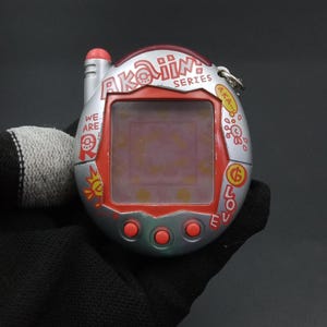 Puede incluir: Un juego electrónico portátil Akaiin Series plateado y rojo. El dispositivo tiene una pequeña pantalla, tres botones rojos y las palabras "WE ARE" y "LOVE". El juego se sostiene en una mano enguantada de negro.