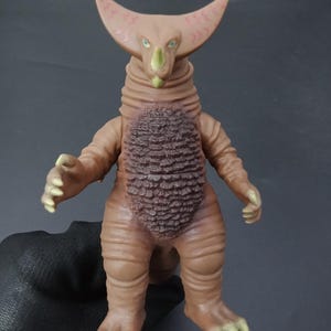 May include: A brown monster action figure with a textured body, large curved horns, and a detailed chest. The figure has a green snout, blue eyes, and yellow claws. The toy is a collectible item.