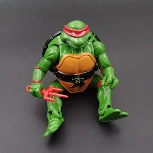 Puede incluir: Una figura de acción verde de Rafael de las Tortugas Ninja. La figura tiene una máscara roja, un caparazón marrón y un cinturón negro con una "R". Sostiene dos armas sai rojas.