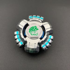 Op de afbeelding: Een zilverkleurige metalen tol met een centrale witte zeshoekige bout voorzien van een groene leeuwengrafiek. Het ronde lichaam heeft vijf gesegmenteerde buitenste secties met heldere cyaanblauwe accenten tussen de metalen ribbels.