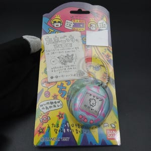 May include: A vintage Tamagotchi virtual pet toy, still in its original packaging. The packaging is colorful with Japanese text and illustrations. The Tamagotchi itself is blue and pink, with a small screen displaying a digital pet. The package reads "BANDAI 1997".