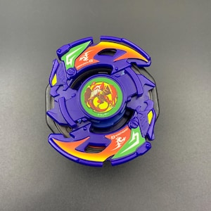 May include: A purple spinning top toy featuring a central circular emblem with a red phoenix graphic on a green background. The outer ring has orange and green accents with white text and symbols, including the word Dranzer G in white lettering.