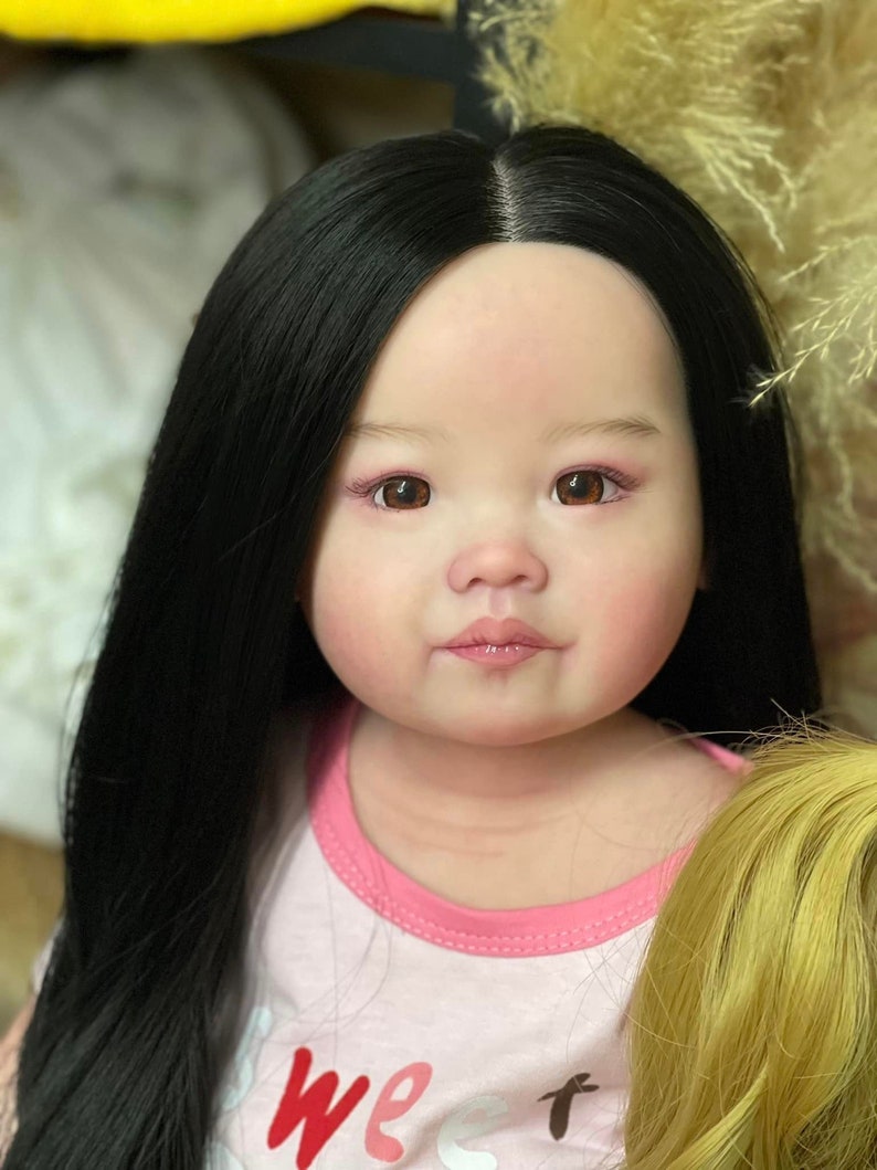 以下が含まれることがあります： 長い黒髪の赤ちゃん人形のクローズアップ。ピンクのシャツを着ており、シャツには「we」という文字が印刷されています。人形は茶色の目をしており、リアルな顔を描いています。