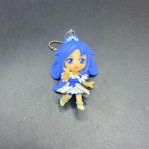Pode incluir: Uma estatueta azul e branca em estilo anime com um chaveiro. A estatueta tem cabelo azul, um vestido branco e azul e detalhes amarelos. É um pequeno item colecionável detalhado, provavelmente um personagem de uma série animada japonesa.