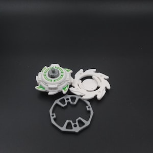 May include: A white and green toy spinning top with a silver central post, a white circular piece with curved edges, and a grey ring with geometric cutouts. The pieces are on a dark surface.
