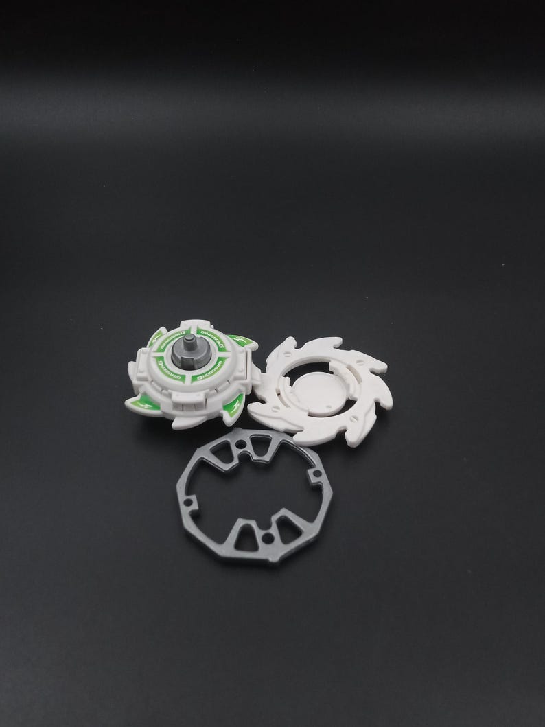 May include: A white and green toy spinning top with a silver central post, a white circular piece with curved edges, and a grey ring with geometric cutouts. The pieces are on a dark surface.