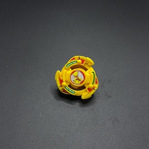 Könnte beinhalten: Gelbe und grüne Plastik-Beyblade mit einem roten und weißen Aufkleber, der einen Cartoon-Vogel mit dem Text "Beyblade" auf dem Aufkleber zeigt.