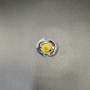 Puede incluir: Un juguete giratorio plateado y negro con un centro amarillo que presenta un gráfico de abeja. El juguete tiene un diseño circular con bordes curvos y está sobre un fondo gris oscuro.