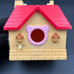 Op de afbeelding: Een beige en rood speelgoedhuis met een rood dak, een rond roze raam en twee eikelvormige versieringen. Het huis heeft een baksteenachtige basis en twee kleine schoorstenen. Onder het raam is een kleine witte knop zichtbaar.