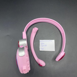 Pode incluir: Um fone de ouvido de brinquedo rosa com detalhes prateados e um design curvo. O fone de ouvido tem um topo arredondado e uma extensão semelhante a um microfone. Um pequeno papel branco com texto está ao lado do fone de ouvido.