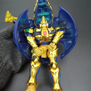 May include: A gold and blue action figure with a robotic insect-like design. The figure has large blue wings, gold accents, and a red center. The toy is posed upright, showcasing its intricate details and design.