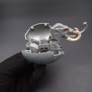 May include: A grey plastic toy mammoth, with an egg-shaped shell that opens. The mammoth has white tusks and red eyes. The toy is partially open, showing internal mechanisms. The toy is held by a gloved hand.