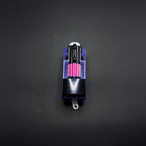 Puede incluir: Un pequeño motor de tren de juguete con un cuerpo morado, frente negro e interior rosa. La parte superior presenta un diseño blanco y negro. El tren tiene un gancho blanco en la parte delantera. El juguete está sobre un fondo negro.