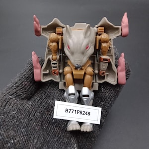 May include: A tan and brown toy robot in a rat-like form with pink accents. The robot has a gray head with red eyes and silver feet. A white label with the text "B771P8248" is attached.