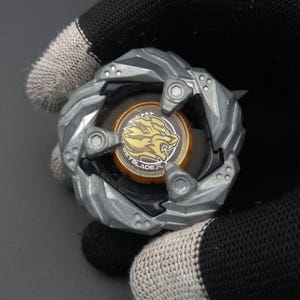 Pu&ograve; includere: Un giocattolo argentato e nero con un centro dorato raffigurante un design a tigre e la scritta "BEYBLADE". Il giocattolo ha una forma circolare con un bordo esterno strutturato e scanalato e tre viti visibili.