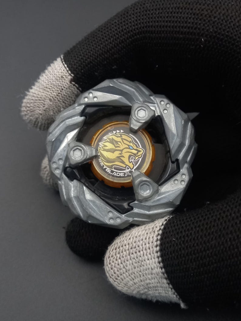 Pu&ograve; includere: Un giocattolo argentato e nero con un centro dorato raffigurante un design a tigre e la scritta "BEYBLADE". Il giocattolo ha una forma circolare con un bordo esterno strutturato e scanalato e tre viti visibili.