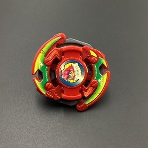 Pode incluir: Beyblade de plástico vermelho, verde e amarelo com um personagem de desenho animado na face. O Beyblade tem um centro preto e uma ponta de metal prateada.