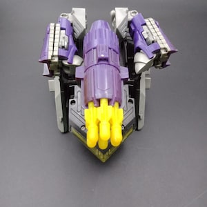 May include: A purple and gray toy robot with yellow accents. The toy has a central purple structure with a cylindrical shape, flanked by articulated arms. The base is a dark gray with yellow protrusions.
