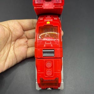 May include: A red toy fire truck with a ladder and emergency lights. The toy has a detailed design with windows, a grill, and silver accents. The toy is held by a hand against a dark background.