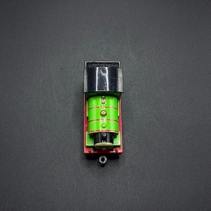 Puede incluir: Un tren de juguete verde y negro con una base roja. El motor tiene una chimenea negra y una cara negra con dos ojos negros y una boca negra.