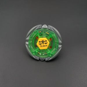 Könnte beinhalten: Ein kreisförmiges Kreiselspielzeug mit einem äußeren Ring aus silberfarbenem Metall und einem inneren Rahmen aus durchscheinendem Grün. In der Mitte befindet sich eine gelbe sechseckige Schraube mit dem schwarzen Waage-Symbol und dem Schriftzug Libra.